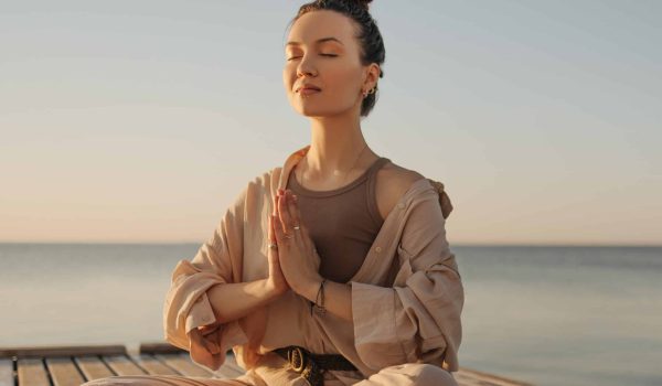 Relaxed,Young,Caucasian,Woman,Sitting,On,Seashore,Practices,Yoga,Without Relaxed young caucasian woman sitting on seashore practices yoga without stress. Model with dark topknot on her head with eyes closed. Cozy beach atmosphere, summer concept.